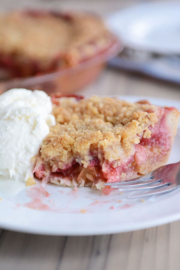 A streusel-topped, berry-filled slice of pie, with one bite taken out, on a white plate, with a scoop of vanilla ice cream next to it, and the rest of the pie in a pie plate in the background.