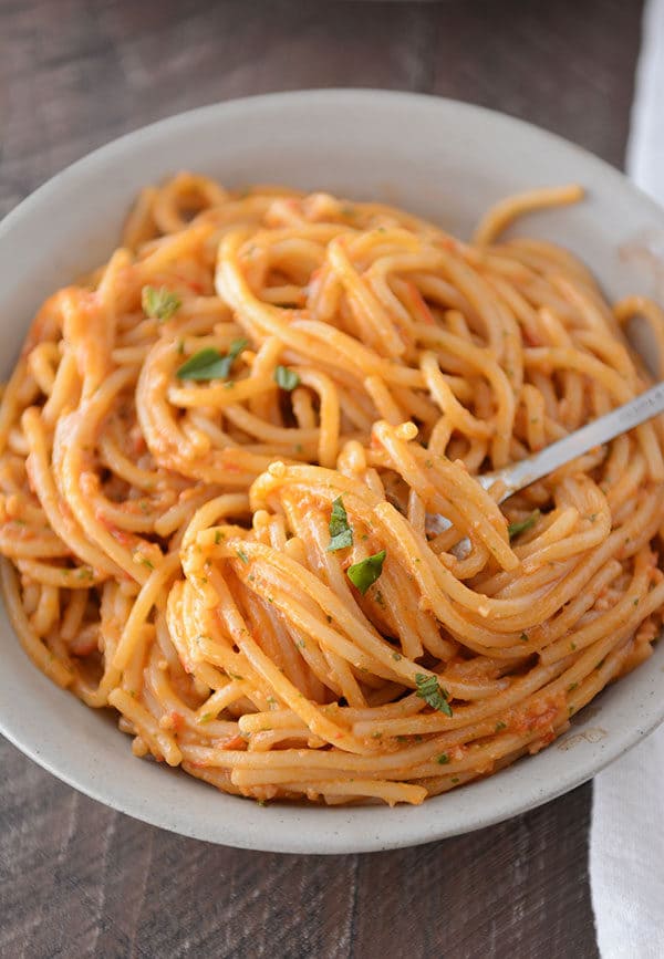 A fork getting a large bite of tomato pesto spaghetti noodles on it from a white bowl of pasta.