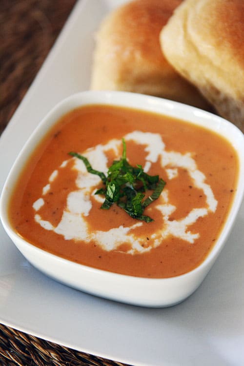 A white bowl of creamy tomato soup with two rolls behind it.