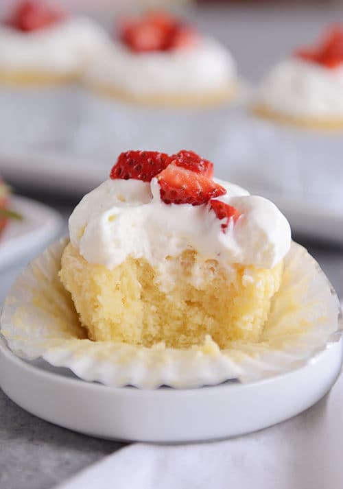 A tres leches cupcake topped with frosting and strawberries with a bite taken out. 