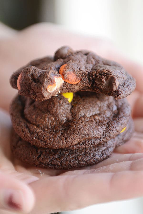 A hand holding a stack of chocolate M&M cookies with a a bite taken out of the top one.