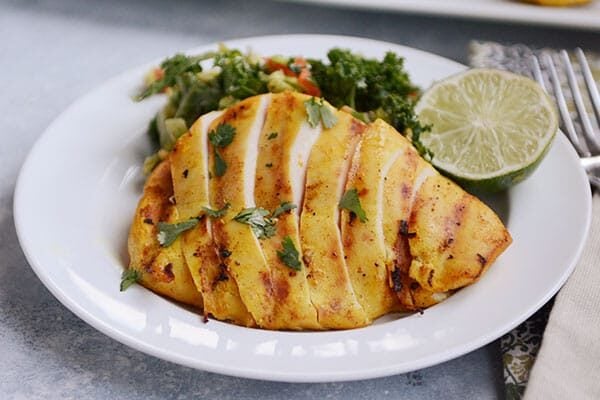 A white plate with sliced grilled chicken and a green salad and slice of lime on the plate.