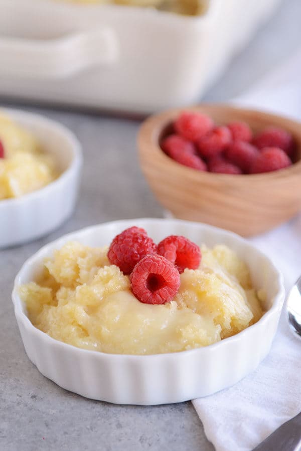A white ramekin full of vanilla pudding cake topped with fresh raspberries a bowl of more raspberries behind it.