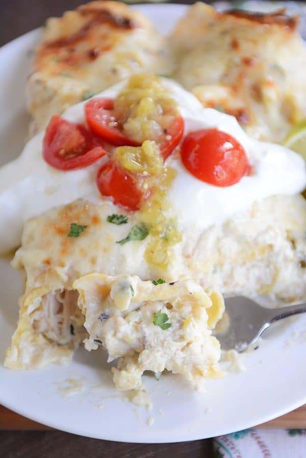 Two baked green chile chicken enchiladas side-by-side on a plate, topped with sour cream and cherry tomatoes and sliced limes on the side.