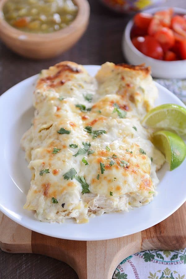 Two baked green chile chicken enchiladas side-by-side on a plate, topped with sour cream and cherry tomatoes and sliced limes on the side.