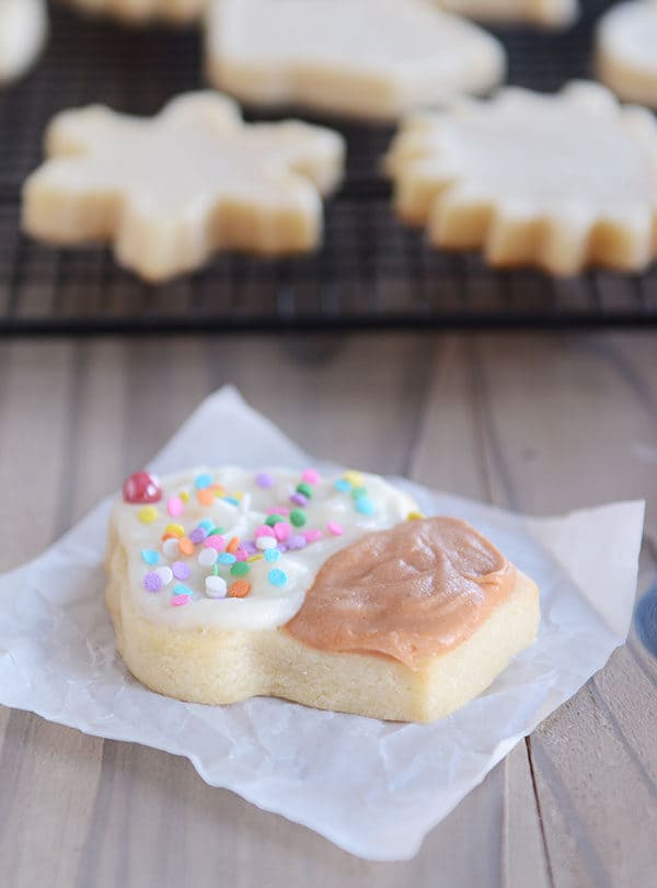 A sugar cookie cut out and decorated like a cupcake on a piece of white parchment paper, with unfrosted sugar cookies on a cooling rack in the background.