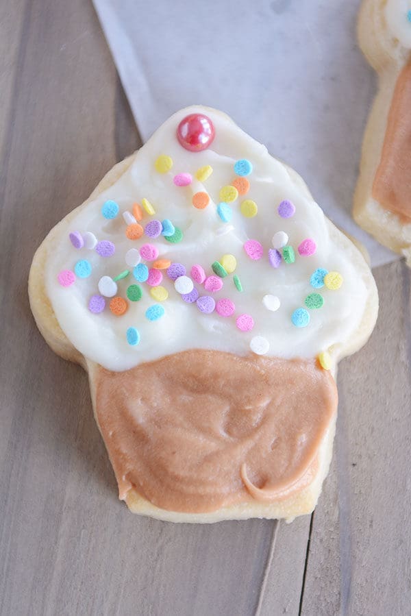 A white sugar cookie decorated like a cupcake with a brown frosting on the bottom, and white frosting and sprinkles on the top of the cookie.