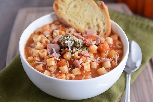 A white bowl minestrone soup with a piece of garlic bread on the side.