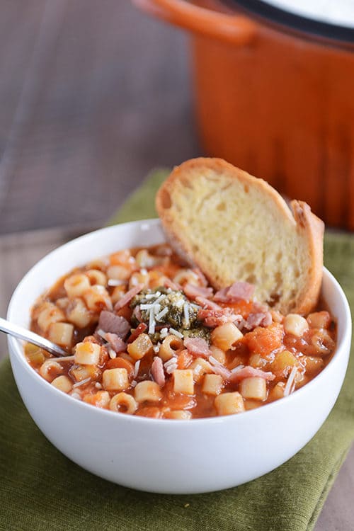 A white bowl full of winter minestrone soup with a piece of garlic bread on the side.