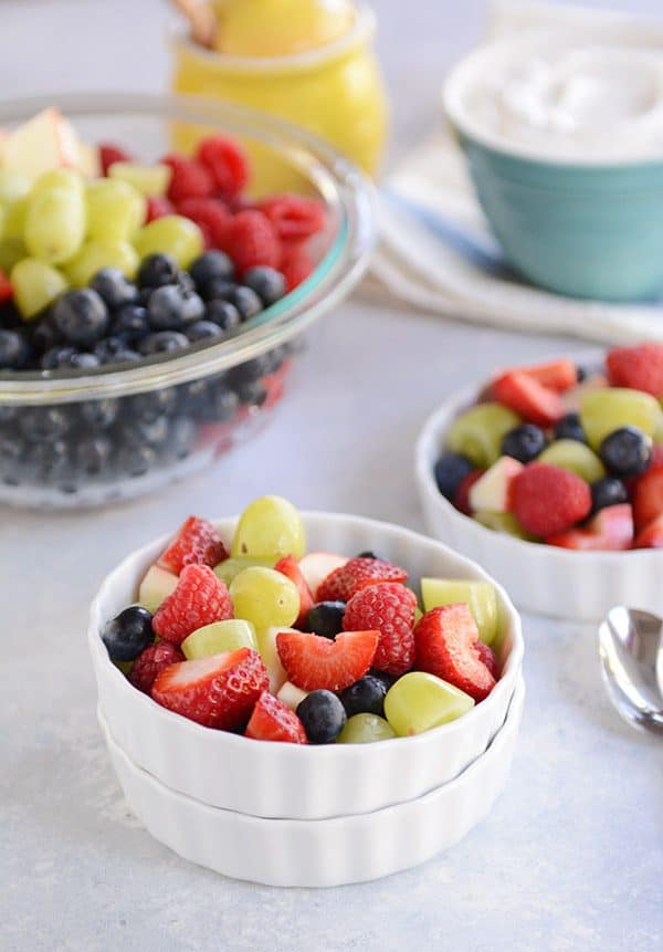 Two white ramekins full of fresh fruit and a large glass bowl full of fruit salad.