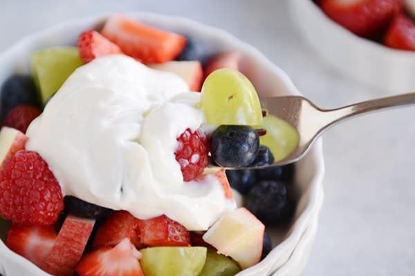 A fork taking a bite of yogurt-topped fruit salad out of a white ramekin.