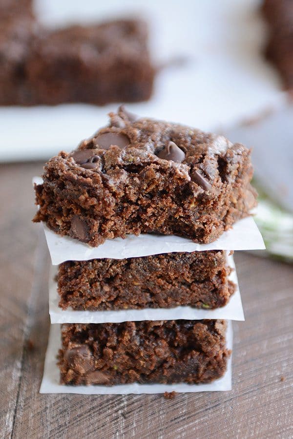 Three chocolate chip zucchini brownies stacked on top of each other and layered between sheets of parchment paper.