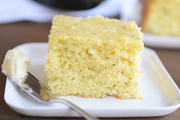 A thick piece of zucchini cornbread on a white plate with a knife with butter on the side.