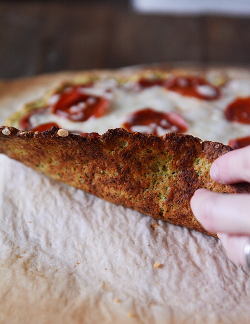 A hand lifting up a pizza to show the bottom of a cooked zucchini crust.