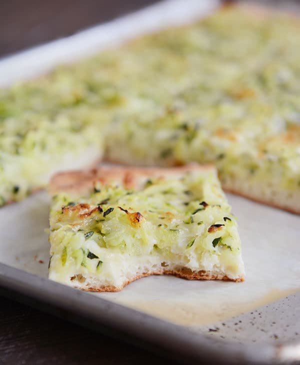 A cookie sheet of zucchini-topped pizza with some pieces cut out and a bite taken out of one piece.
