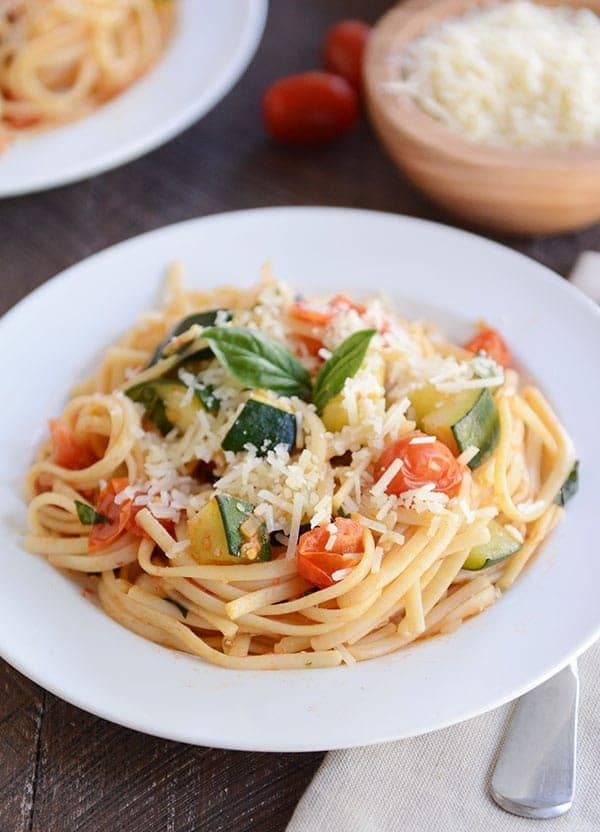 A white plate of zucchini tomato pasta topped with parmesan cheese and basil leaves on a white plate.
