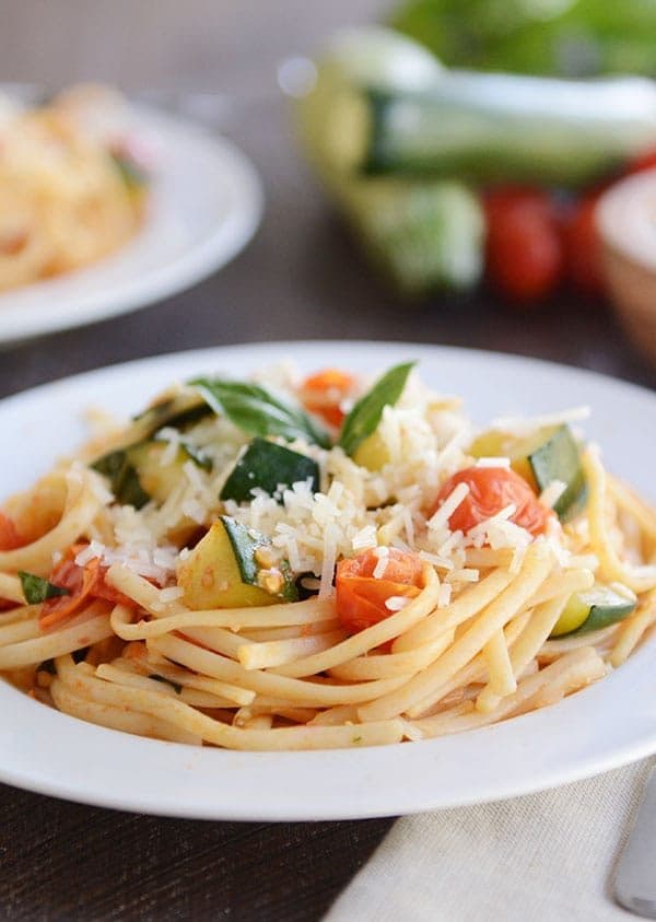 A white plate of pasta with chopped zucchini and tomato on top.
