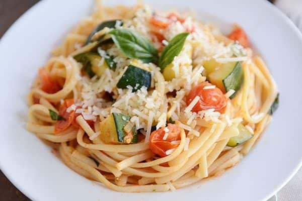 A white plate of zucchini and tomato pasta topped with two basil leaves.